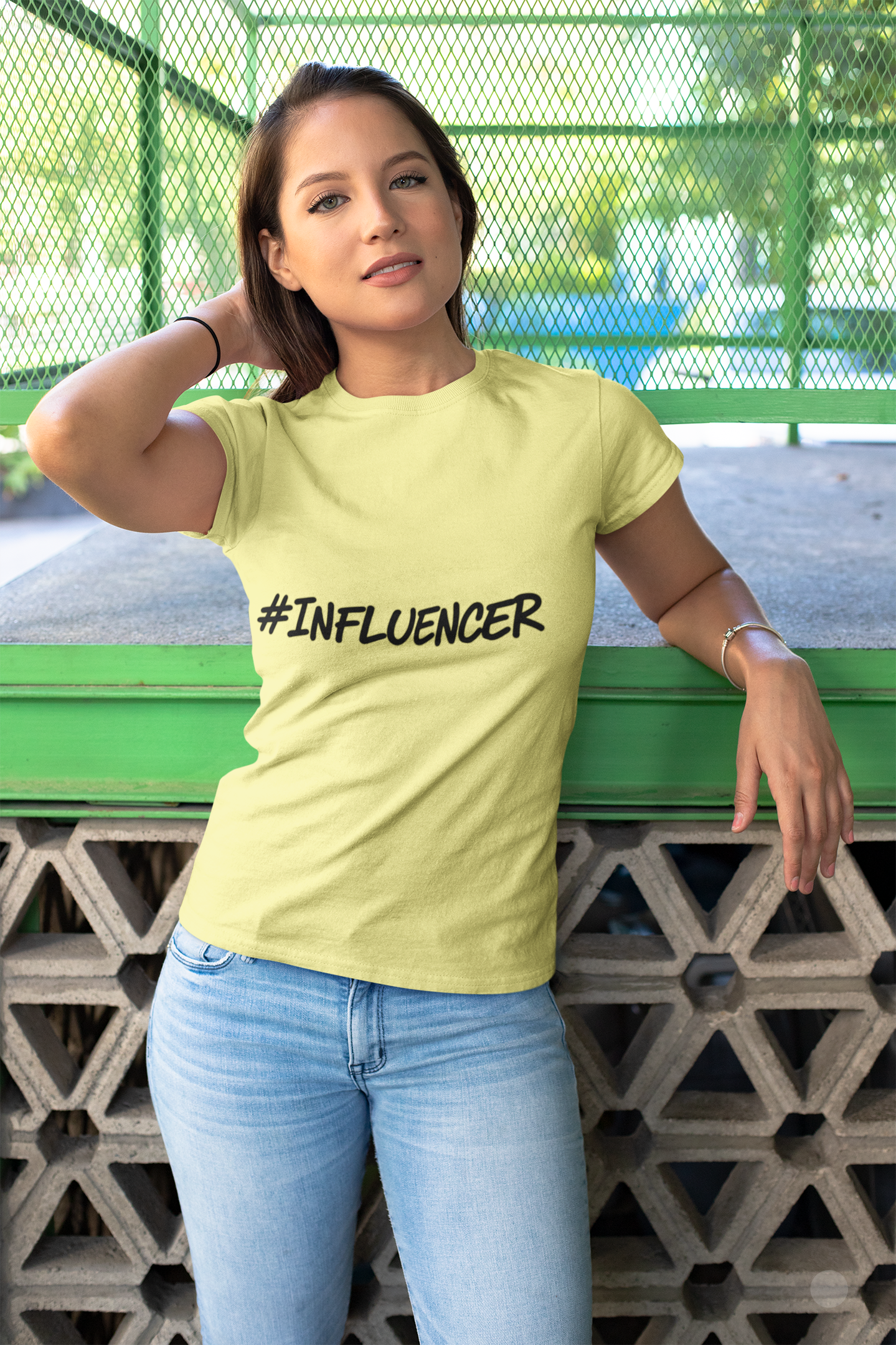 Woman wearing a yellow '#INFLUENCER' t-shirt standing against a green metal fence.